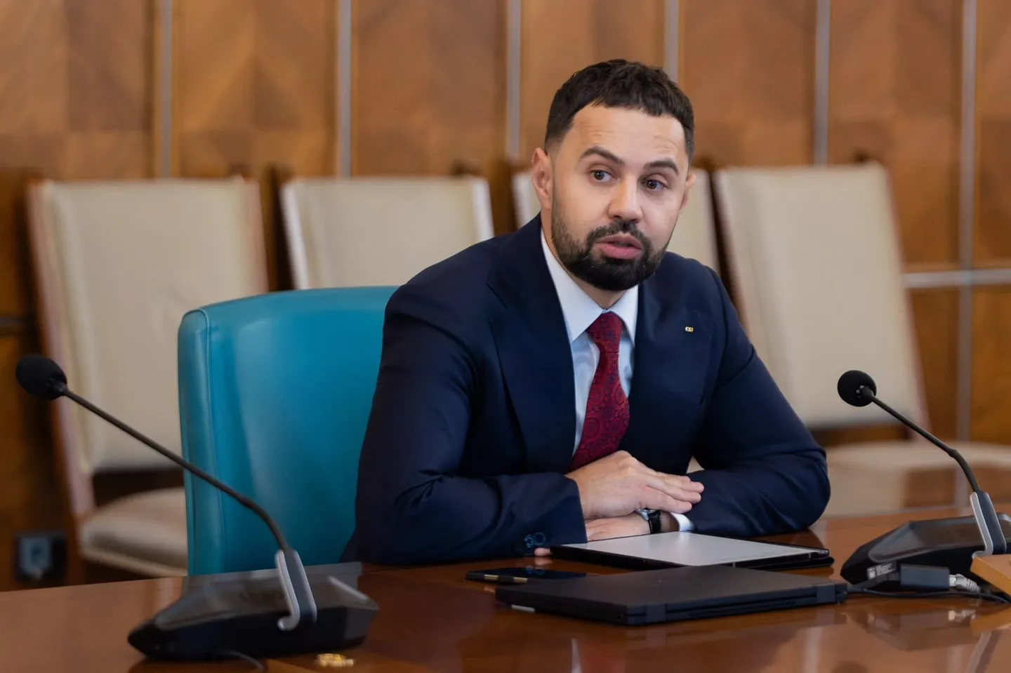 Un bărbat cu barbă, îmbrăcat în costum închis la culoare și cravată roșie, stă la masă între două microfoane.