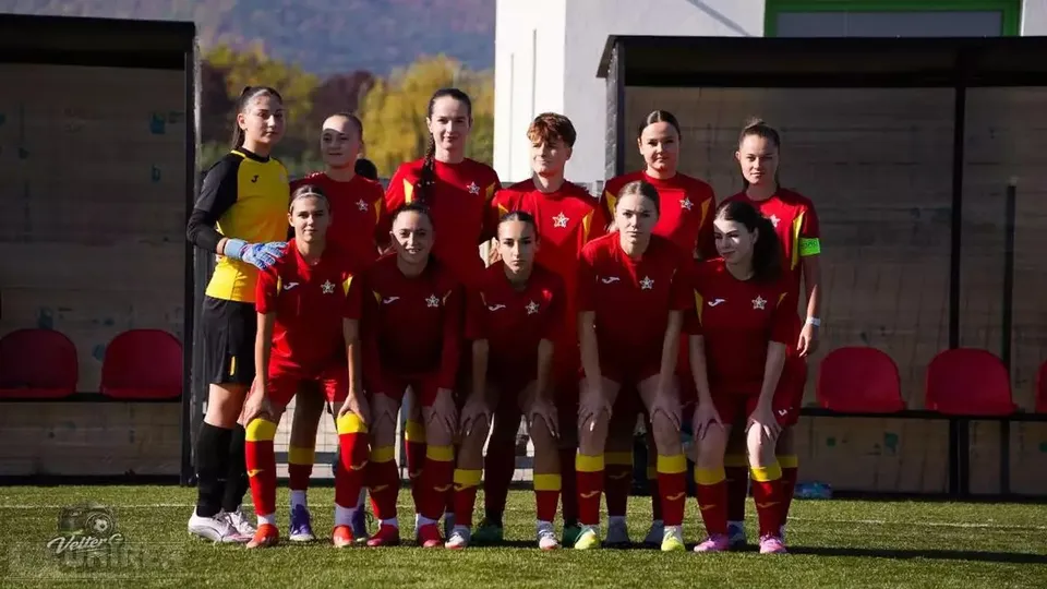 Un grup de sportive în echipamente roșii și negre pozează pe iarbă, în fața unor bănci acoperite cu scaune roșii.
