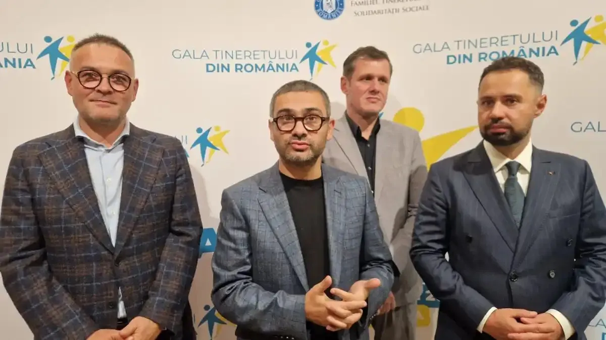 Patru bărbați în sacouri stau în fața unui panou cu text și simboluri colorate; unul este parțial ascuns în spatele celorlalți.