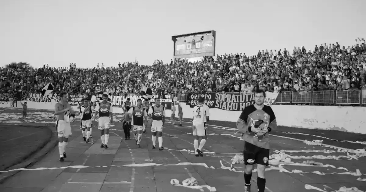 Mai mulți bărbați în echipament sportiv merg pe o pistă, în fața unei tribune pline; bărbatul din prim-plan ține o minge.