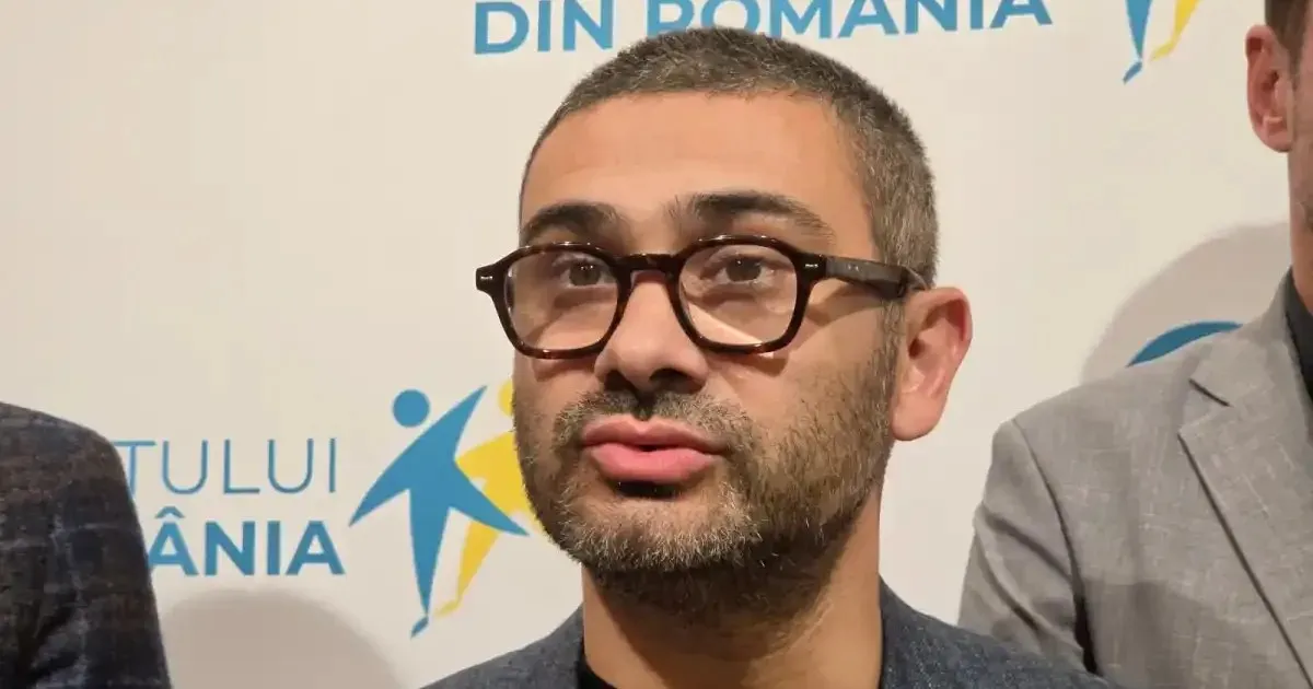 Un bărbat cu păr scurt, barbă și ochelari cu rame închise apare în prim-plan, lângă două persoane parțial vizibile.