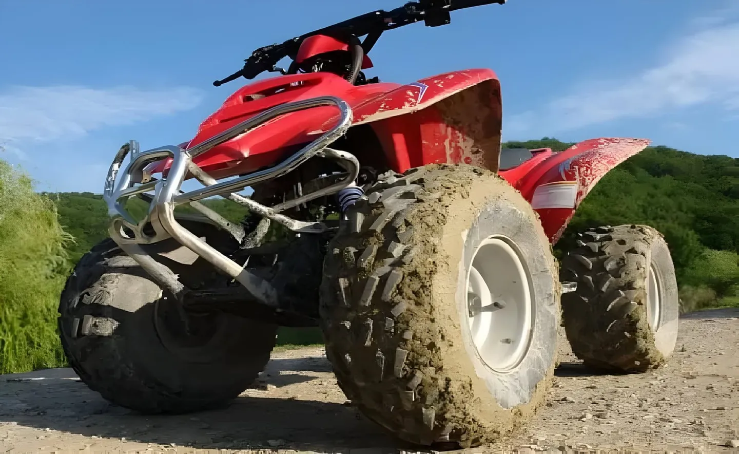 Un ATV roșu, cu anvelope acoperite de noroi, este fotografiat de aproape pe un drum pietruit, lângă vegetație verde.