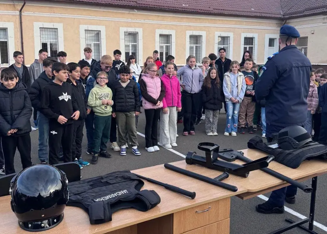 Un bărbat în uniformă stă în fața unui grup de copii, iar pe mesele din prim-plan se află o cască, veste negre și bastoane.