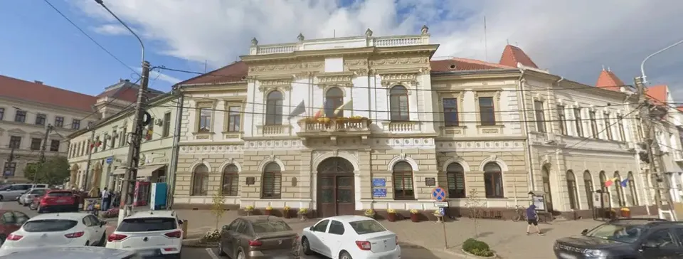 O clădire cu fațadă ornamentată se află în spatele mai multor autoturisme parcate, sub un cer parțial înnorat.