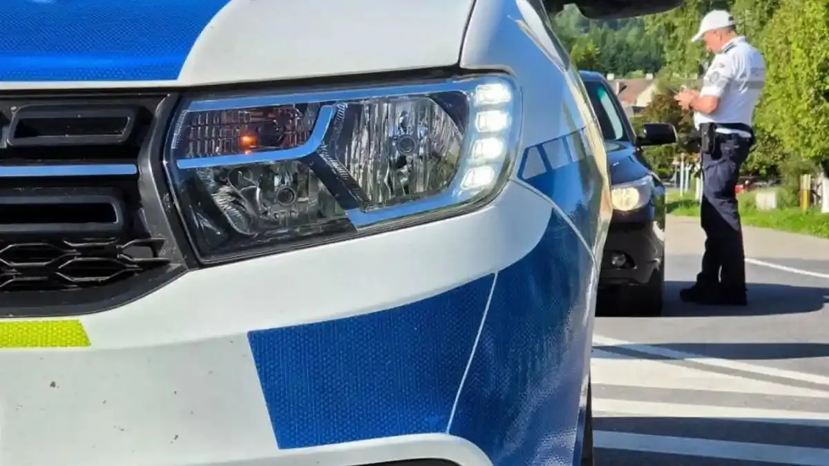 În prim-plan se vede partea frontală a unei autospeciale alb-albastre; în fundal, un bărbat în uniformă deschisă la culoare stă lângă un autoturism închis la culoare.