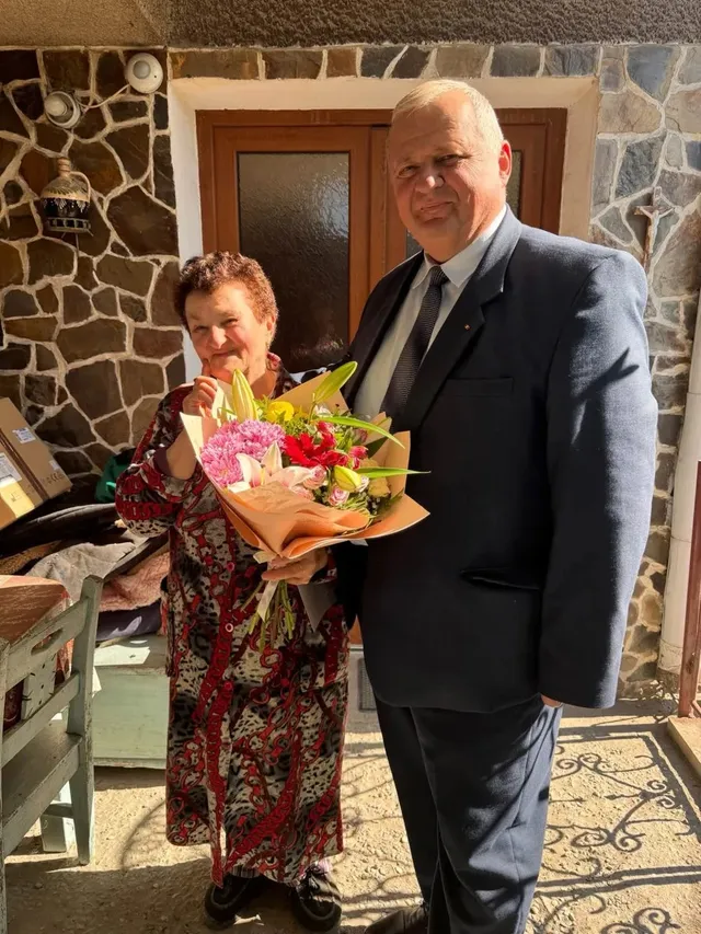 O femeie în rochie colorată ține un buchet de flori lângă un bărbat în costum, în fața unei uși și a unui perete din piatră.