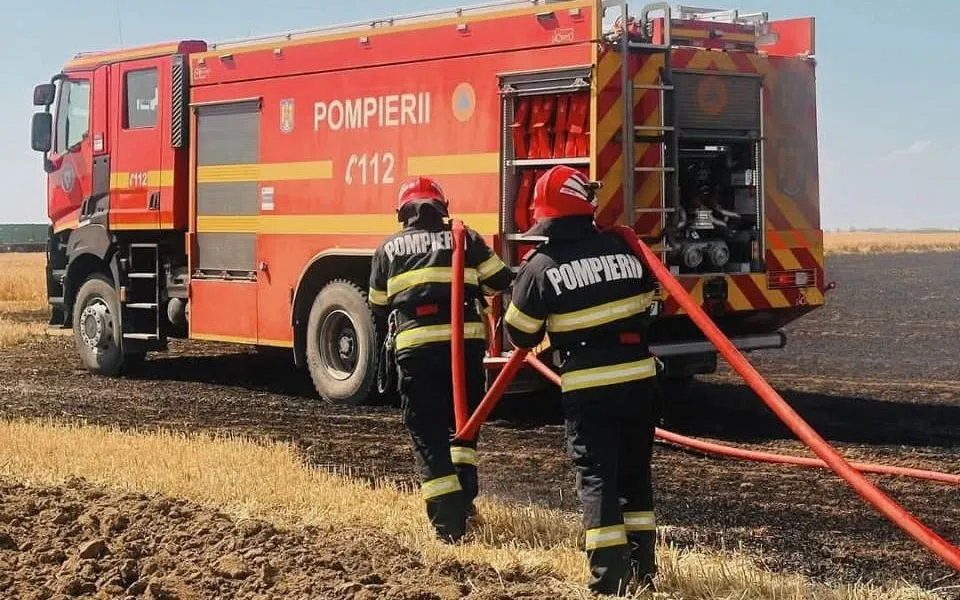 O autospecială roșie și doi pompieri în uniforme negre țin furtunuri roșii pe un câmp cu vegetație arsă și brazde în prim-plan.