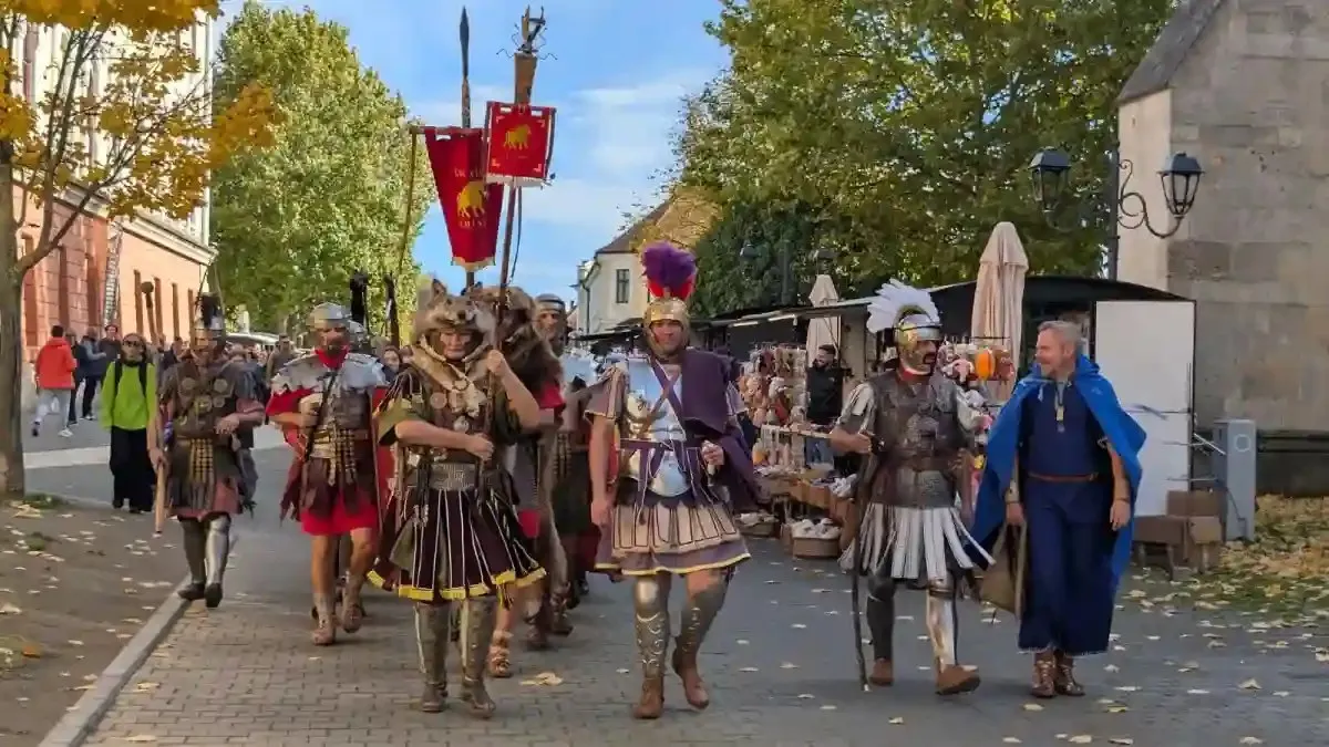Mai multe persoane în costume antice, cu coifuri, scuturi și sulițe, merg pe o alee; în spate se văd steaguri și copaci.