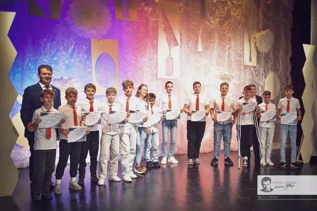 Un bărbat și mai mulți tineri îmbrăcați predominant în alb stau pe o scenă, ținând diplome și purtând medalii.