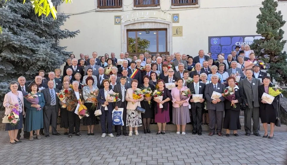 Un grup numeros de adulți vârstnici, îmbrăcați elegant, stă în fața unei clădiri, iar mai multe persoane țin flori și diplome.