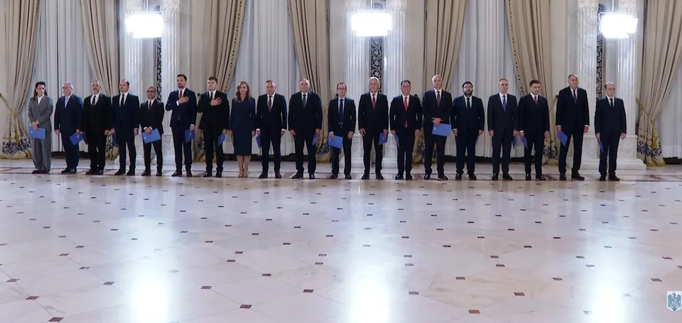 Un grup de persoane în costume închise la culoare stă în șir într-o sală cu draperii, candelabre și pardoseală deschisă la culoare.