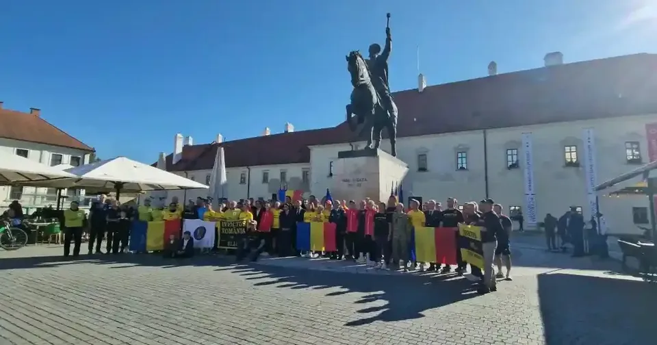 Un grup de persoane ține bannere și steaguri în fața unei statui ecvestre, lângă o clădire albă și umbrele deschise.