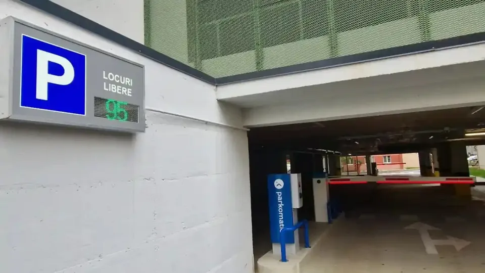 La intrarea într-o parcare acoperită se văd un indicator albastru cu litera P, un afișaj pentru locuri libere și o barieră coborâtă.