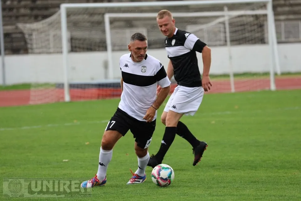 Doi bărbați în echipament alb-negru aleargă pe un teren verde, lângă o minge de fotbal, cu o poartă în fundal.