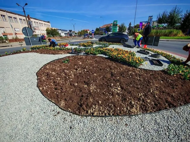 O zonă ovală cu pământ este înconjurată de pietriș deschis la culoare și flori; câteva persoane sunt lângă un drum, iar o mașină se află în apropiere.