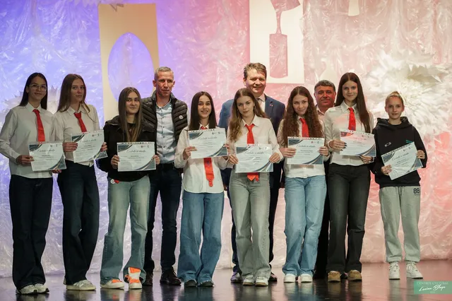Un grup de tineri și adulți stă pe o scenă, iar majoritatea țin diplome, în fața unui fundal iluminat.