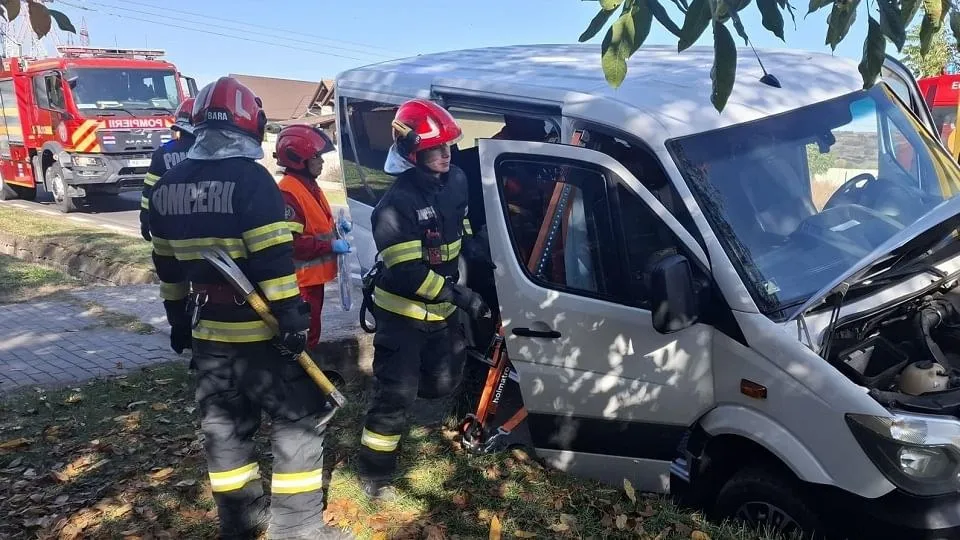 Trei persoane cu căști și echipament de intervenție stau lângă o autoutilitară albă avariată în față; în fundal se vede o autospecială roșie.