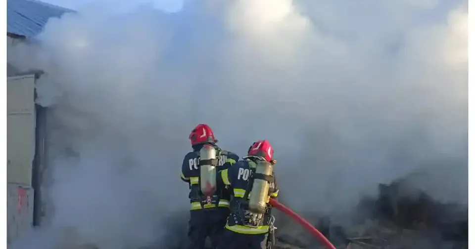 Doi pompieri cu căști roșii și echipament negru stau lângă o clădire, în fața unui nor dens de fum; lângă ei se vede un furtun roșu.