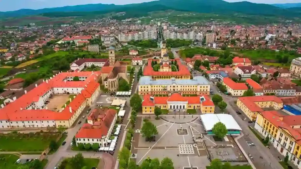 Vedere aeriană cu clădiri cu acoperișuri roșii, o piață pavată în prim-plan, străzi, copaci și dealuri în depărtare.
