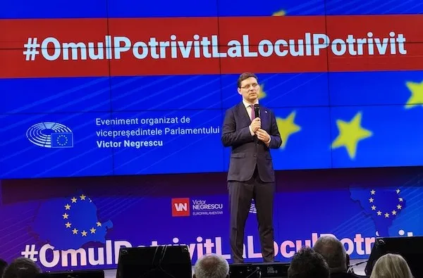 Un bărbat în costum închis la culoare stă pe scenă cu un microfon, în fața unui ecran albastru cu text și stele galbene.