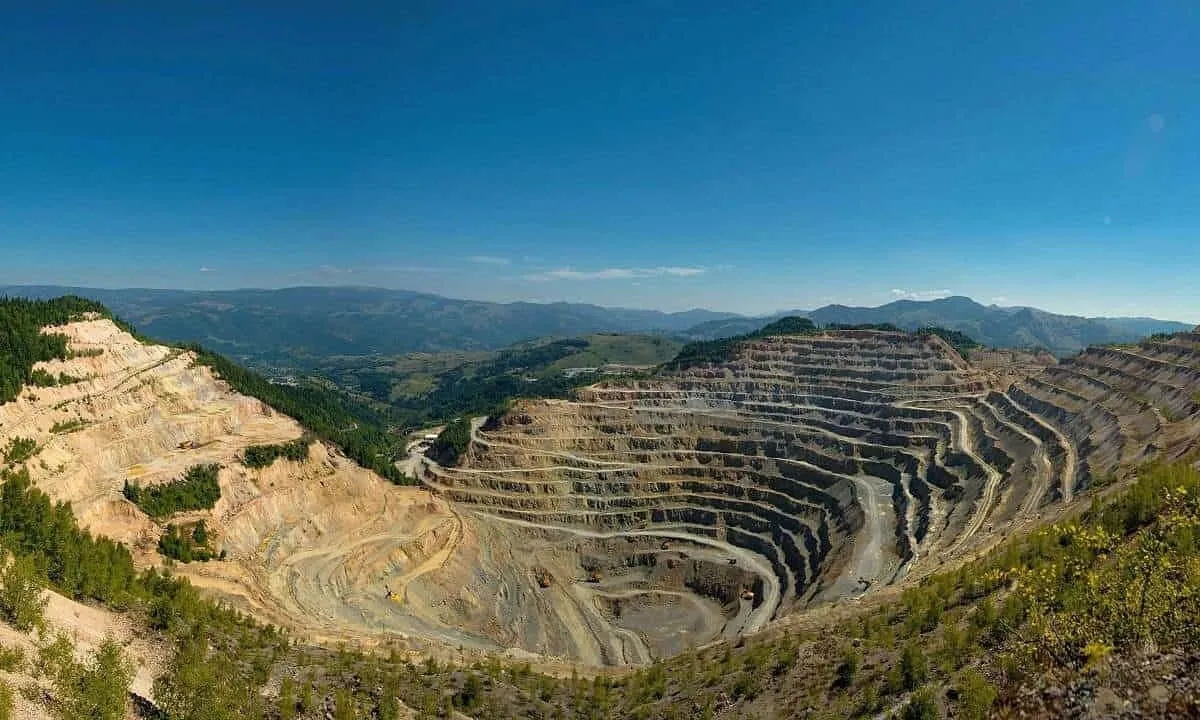 O excavație largă, cu trepte concentrice de pământ și rocă, este înconjurată de versanți împăduriți sub un cer albastru.