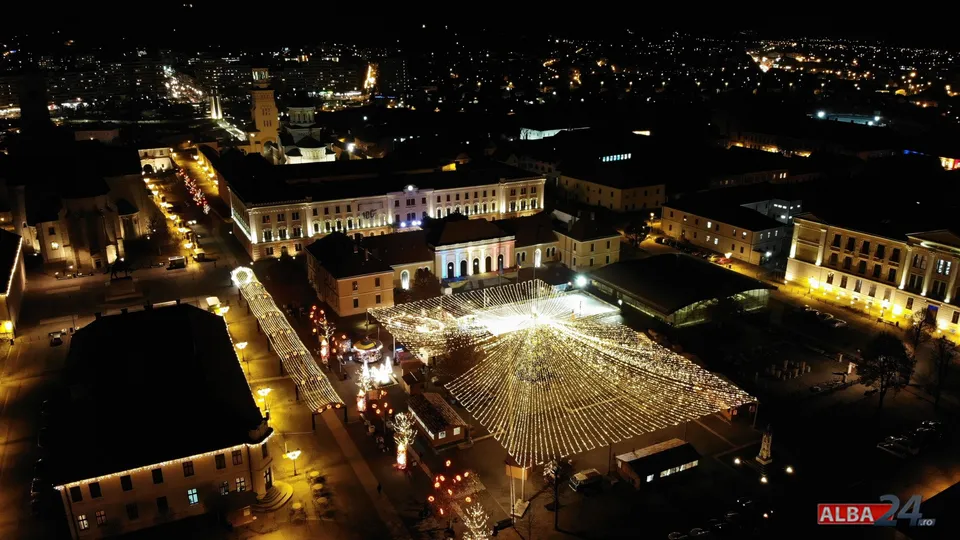Vedere nocturnă de sus asupra unei piețe cu șiruri de lumini, construcții mici, clădiri luminate și străzi în jur.