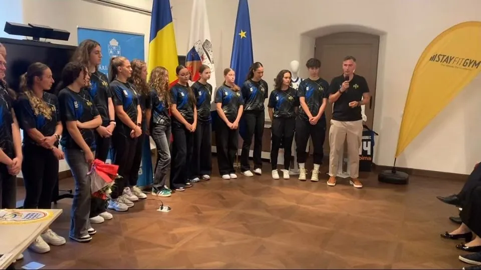 Un grup de tineri în tricouri albastre stă aliniat într-o sală, lângă două steaguri, un bărbat și un banner galben.