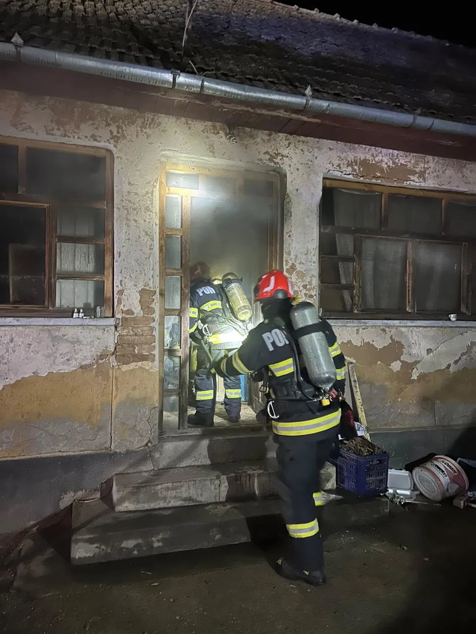 Doi pompieri cu echipamente reflectorizante și butelii pe spate stau la intrarea unei clădiri cu tencuială deteriorată.