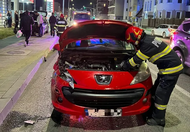 O mașină roșie are capota ridicată și partea frontală avariată; o persoană în echipament reflectorizant stă aplecată lângă motor, iar alte persoane sunt în fundal.