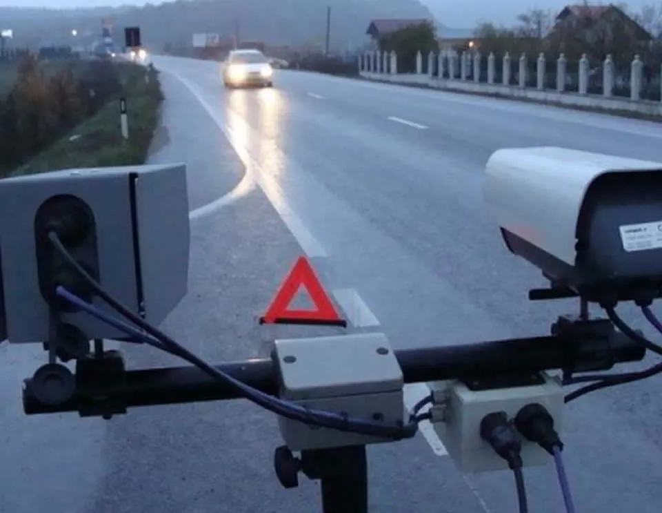 Pe un drum umed se apropie o mașină cu farurile aprinse; în prim-plan se văd un triunghi roșu și aparate montate pe un suport.