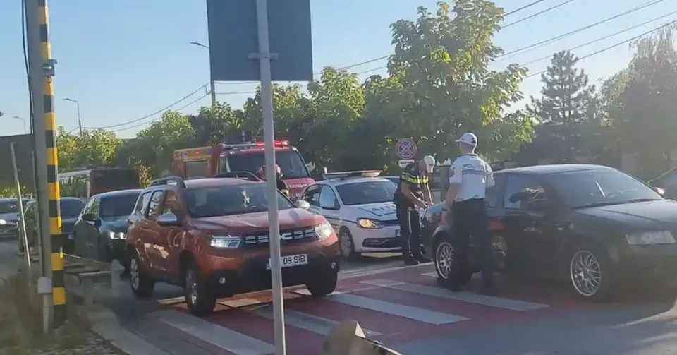 Două autoturisme se află lângă o trecere de pietoni, iar în spatele lor se văd un vehicul roșu și persoane în veste reflectorizante.