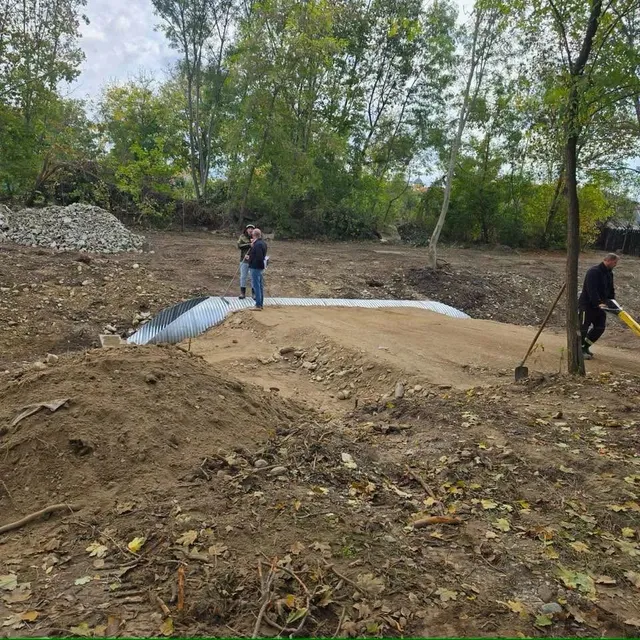O suprafață de pământ nivelat și un tub ondulat se află între grămezi de pământ, cu copaci în spate și doi oameni în picioare.