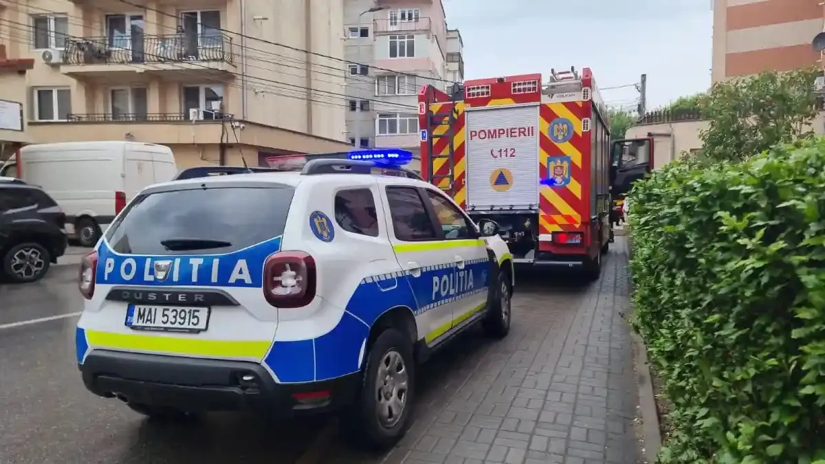 O mașină alb-albastră cu inscripția «POLIȚIA» se află pe carosabil, în spatele unui vehicul roșu cu inscripția «POMPIERII», lângă un trotuar.
