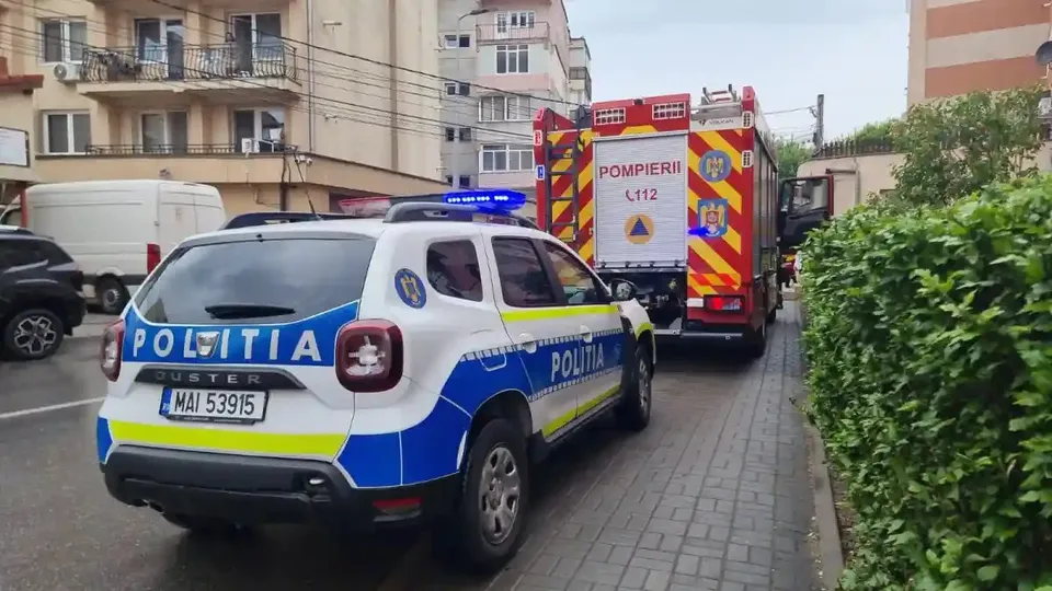 O mașină alb-albastră cu inscripția «POLIȚIA» se află pe carosabil, în spatele unui vehicul roșu cu inscripția «POMPIERII», lângă un trotuar.