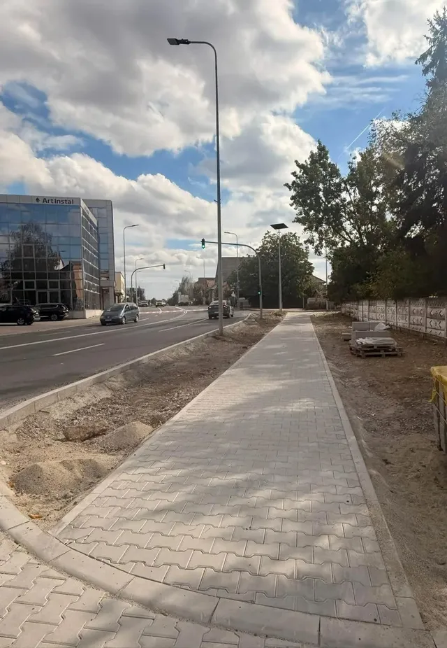 Un trotuar pavat cu dale gri se întinde lângă un drum, cu stâlpi de iluminat, copaci și clădiri în jur.