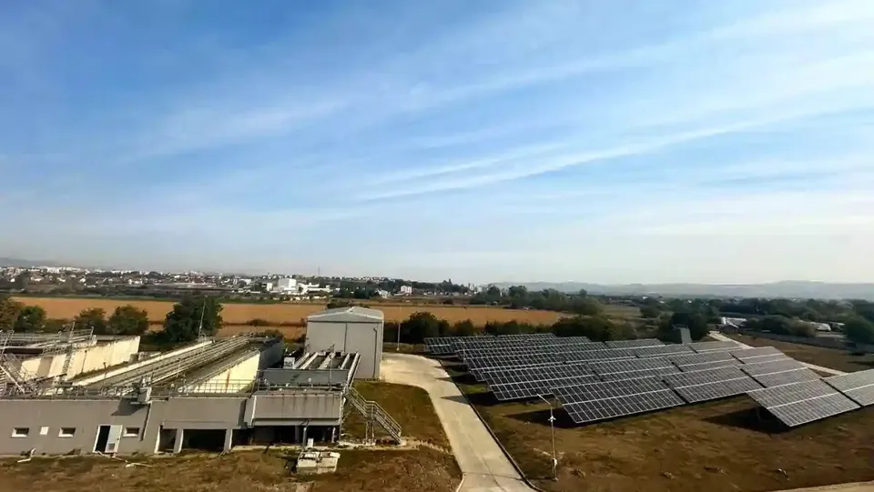 Panouri fotovoltaice sunt dispuse în rânduri pe terenul din dreapta; în stânga se văd clădiri industriale, iar între ele trece un drum.