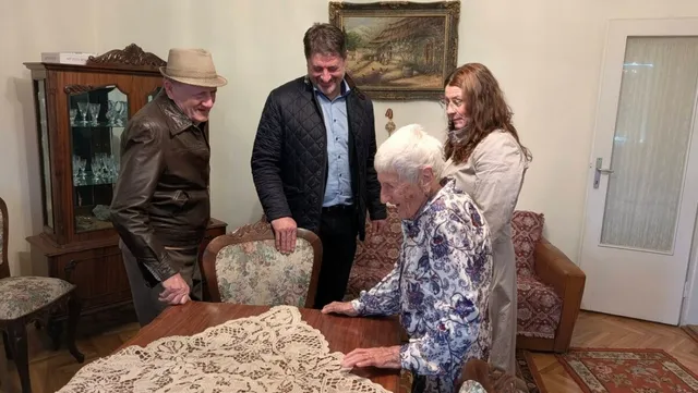 O femeie cu păr alb stă lângă o masă acoperită cu o față de masă croșetată, iar trei adulți stau în jurul ei.