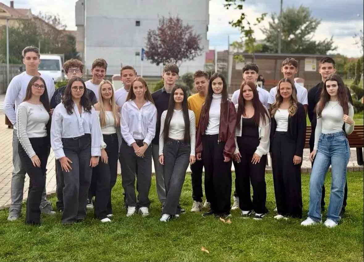Un grup de tineri stă pe iarbă, în aer liber, purtând cămăși, bluze, pantaloni și blugi; în spate se văd clădiri și copaci.