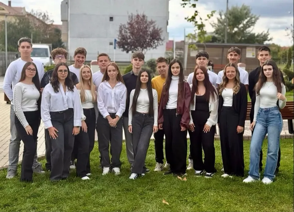 Un grup de tineri stă pe iarbă, în aer liber, purtând cămăși, bluze, pantaloni și blugi; în spate se văd clădiri și copaci.