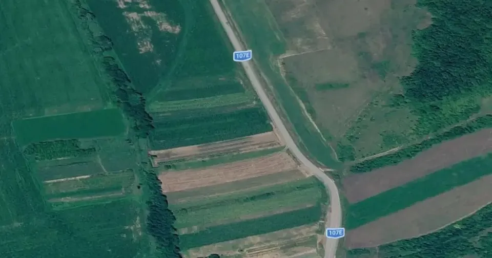 O imagine aeriană arată parcele agricole dreptunghiulare și o șosea care traversează terenurile, cu două indicatoare albastre la marginea drumului.