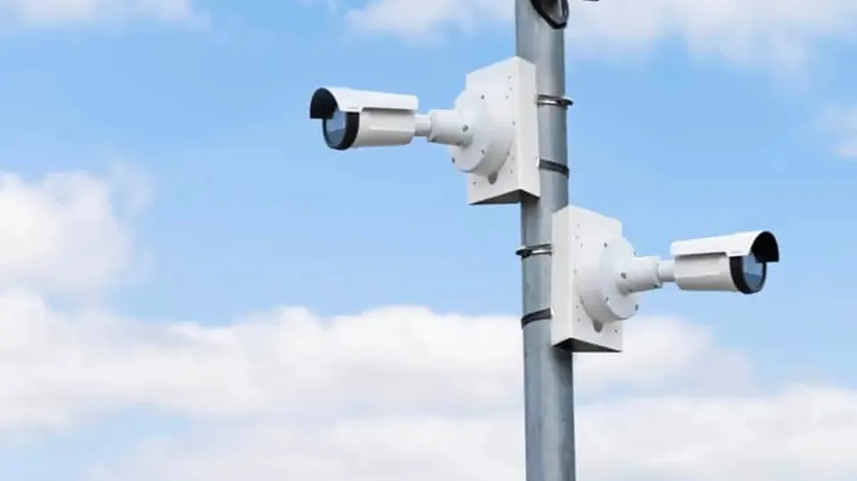 Două camere albe de supraveghere sunt montate pe un stâlp metalic, orientate în direcții opuse, pe fundalul unui cer cu nori.