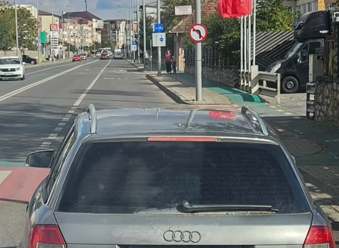 În prim-plan se vede spatele unui autoturism gri, iar pe stradă apar alte vehicule, câțiva pietoni și indicatoare rutiere.