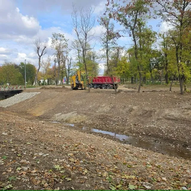 Pe un teren cu pietriș, printre copaci, se văd un excavator galben, un camion roșu și șanțuri cu apă.