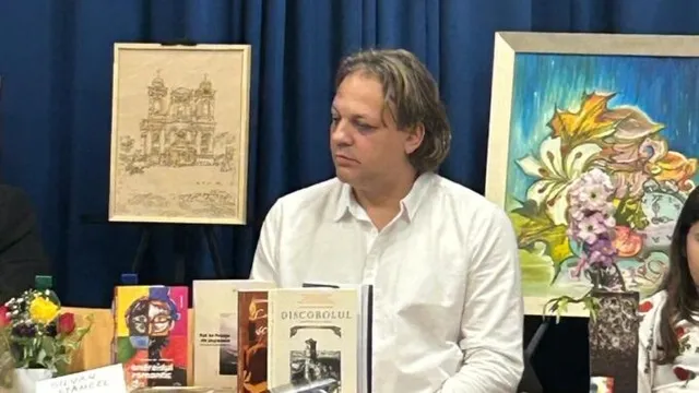 Un bărbat în cămașă albă stă în spatele mai multor publicații expuse; în fundal sunt două tablouri, iar în dreapta apare parțial o persoană.