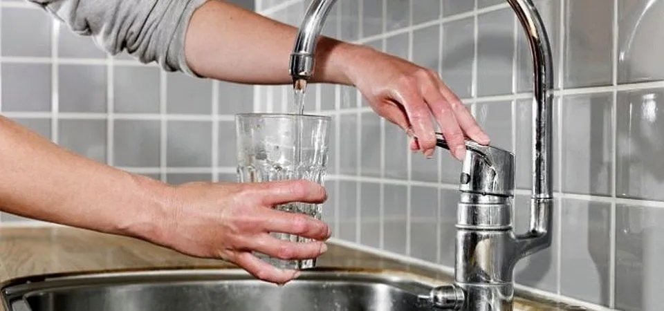 Două mâini țin un pahar sub un robinet metalic din care curge un fir subțire de apă, deasupra unei chiuvete.