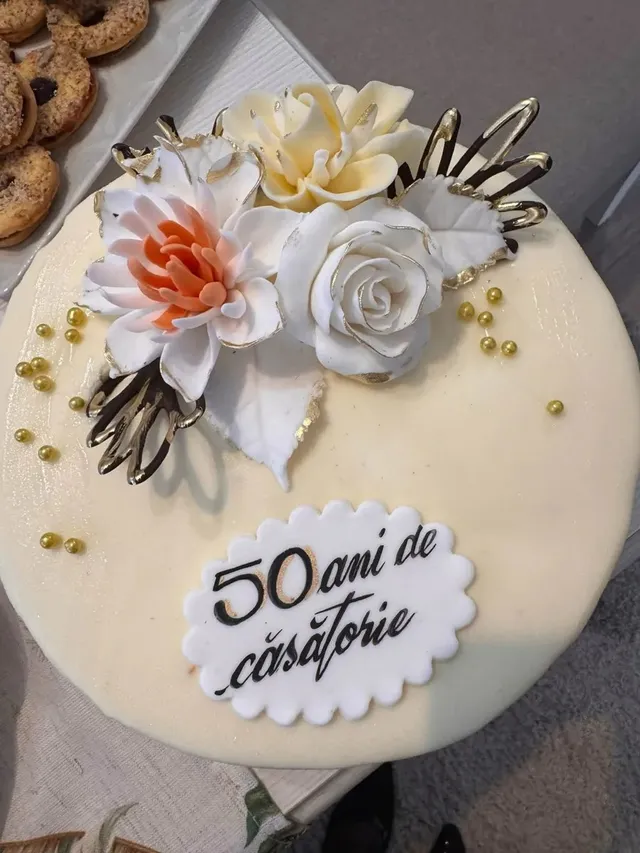 Un tort rotund, alb, decorat cu flori albe și roz, ornamente aurii și o inscripție neagră pe o plăcuță albă.