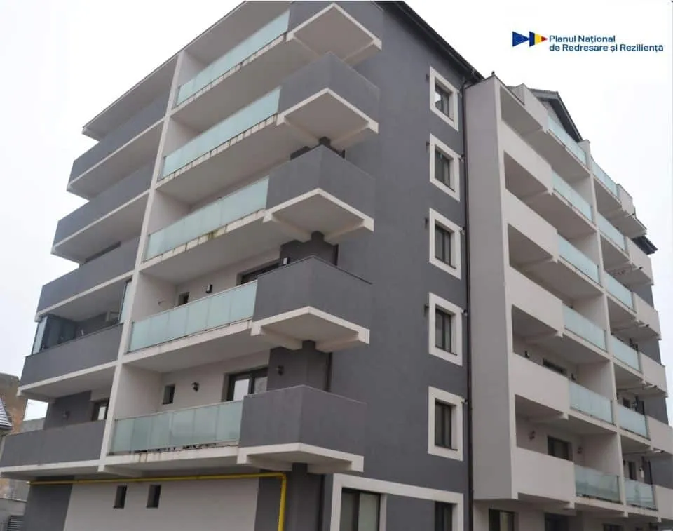 Un bloc cu mai multe etaje are fațade gri și albe, balcoane suprapuse cu balustrade din sticlă și ferestre dreptunghiulare.