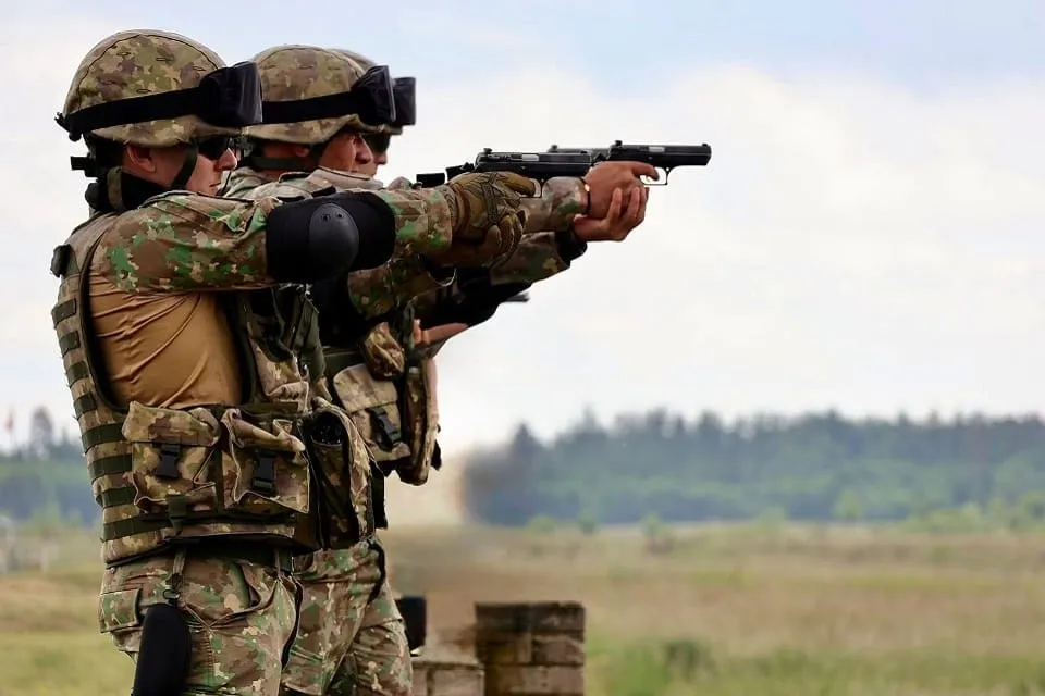 Două persoane în uniforme de camuflaj și căști stau una în spatele celeilalte; cea din față ține un pistol îndreptat spre dreapta.