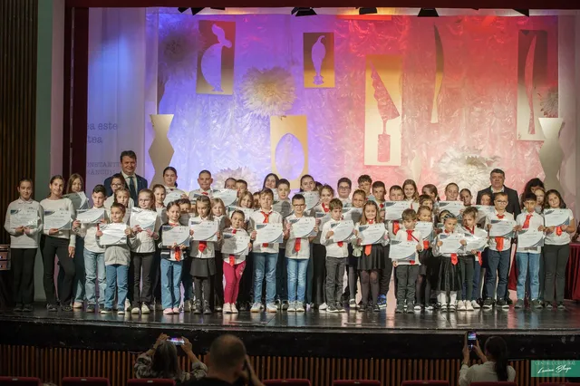 Un grup numeros de copii și câțiva adulți stau pe o scenă, ținând diplome și trandafiri, în fața unui decor colorat.