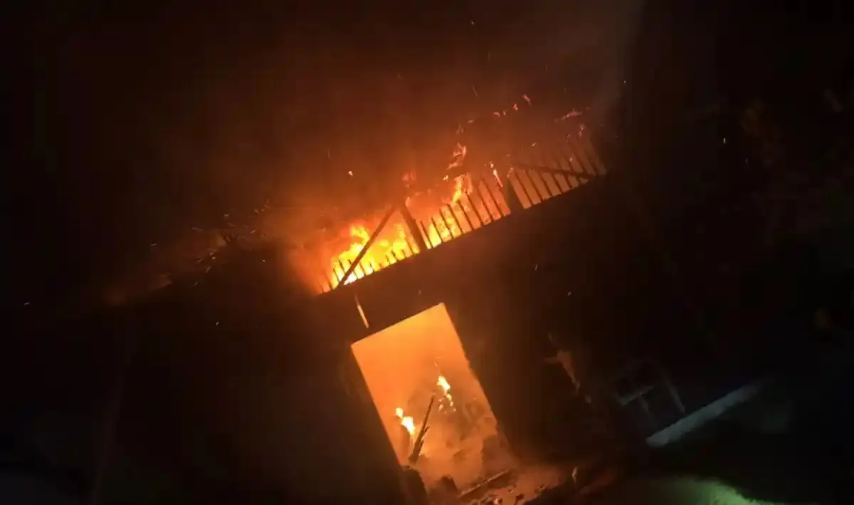 Flăcări și fum se ridică în spatele unui acoperiș înclinat, iar o deschidere dreptunghiulară este luminată de foc.